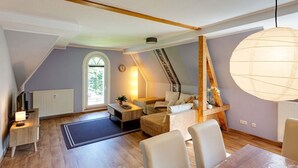 Um quarto de loft acolhedor com teto inclinado, uma janela grande, uma viga de madeira no teto, um lustre e um pequeno espaço de estar com mesa e cadeiras.