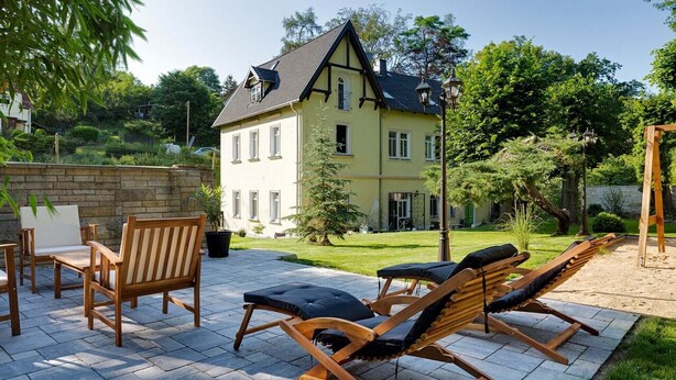 Um pátio com cadeiras de lounge de madeira e piso de pedra, cercado por um jardim e uma casa grande com telhado empena.