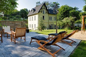 Une terrasse dotée de chaises longues en bois et d’un sol en pierre, entourée d’un jardin et d’une grande maison à toit en pente.