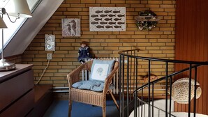 Une pièce chaleureuse avec un mur en briques, un fauteuil en osier, une petite table avec une lampe et un escalier doté d’une rampe.