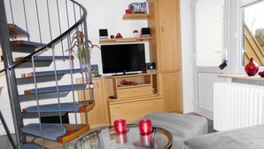 Un salon avec un escalier en colimaçon, une table basse en verre sur laquelle reposent des tasses rouges, un canapé gris et un téléviseur posé sur un meuble en bois.
