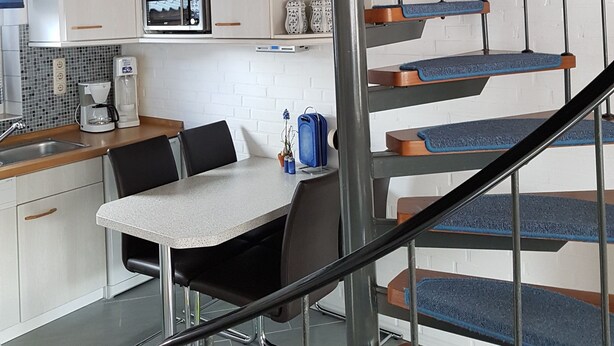 Une cuisine moderne dotée d’un îlot central, d’un coin repas avec une table et des chaises, et d’un escalier en colimaçon dont les marches sont recouvertes d’une moquette bleue.