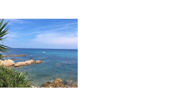 A coastal landscape with clear blue water, rocky formations, and sparse vegetation.