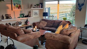 A cozy living room with a brown leather sofa, a wooden coffee table, and a view of a grassy field through large windows. - A farm in a stunning nature. Peace and quiet. Out of tourist traffic. (Dalabyggð)
