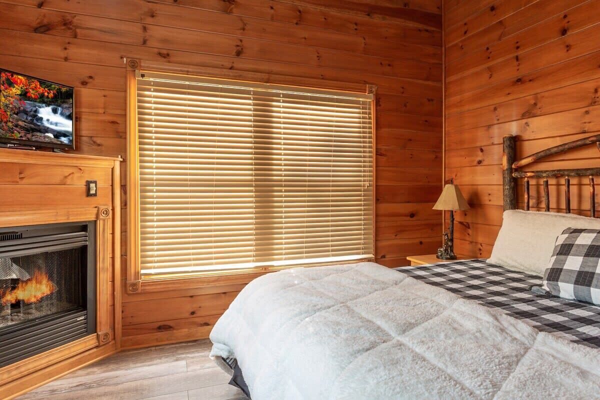 A cozy wooden cabin room with a fireplace, a bed with a checkered blanket, and a large window with blinds.