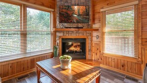 A wooden cabin interior with a stone fireplace, a television mounted above it, and a table with a potted plant.