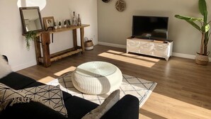 Uma sala de estar com uma mesa consola de madeira, uma televisão sobre um armário branco, uma planta grande em vaso e um tapete estampado.