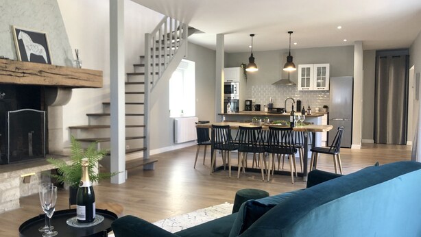Uma sala de estar moderna com um sofá azul, um espaço de jantar com mesa e cadeiras de madeira, uma cozinha com armários brancos e eletrodomésticos de aço inoxidável, e uma escadaria que leva a outro andar.