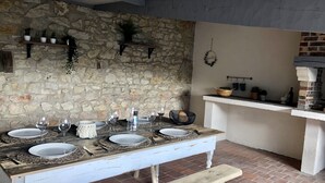 Uma parede de pedra rústica com uma longa mesa de madeira posta para refeição, um banco e um forno de tijolos ao fundo.