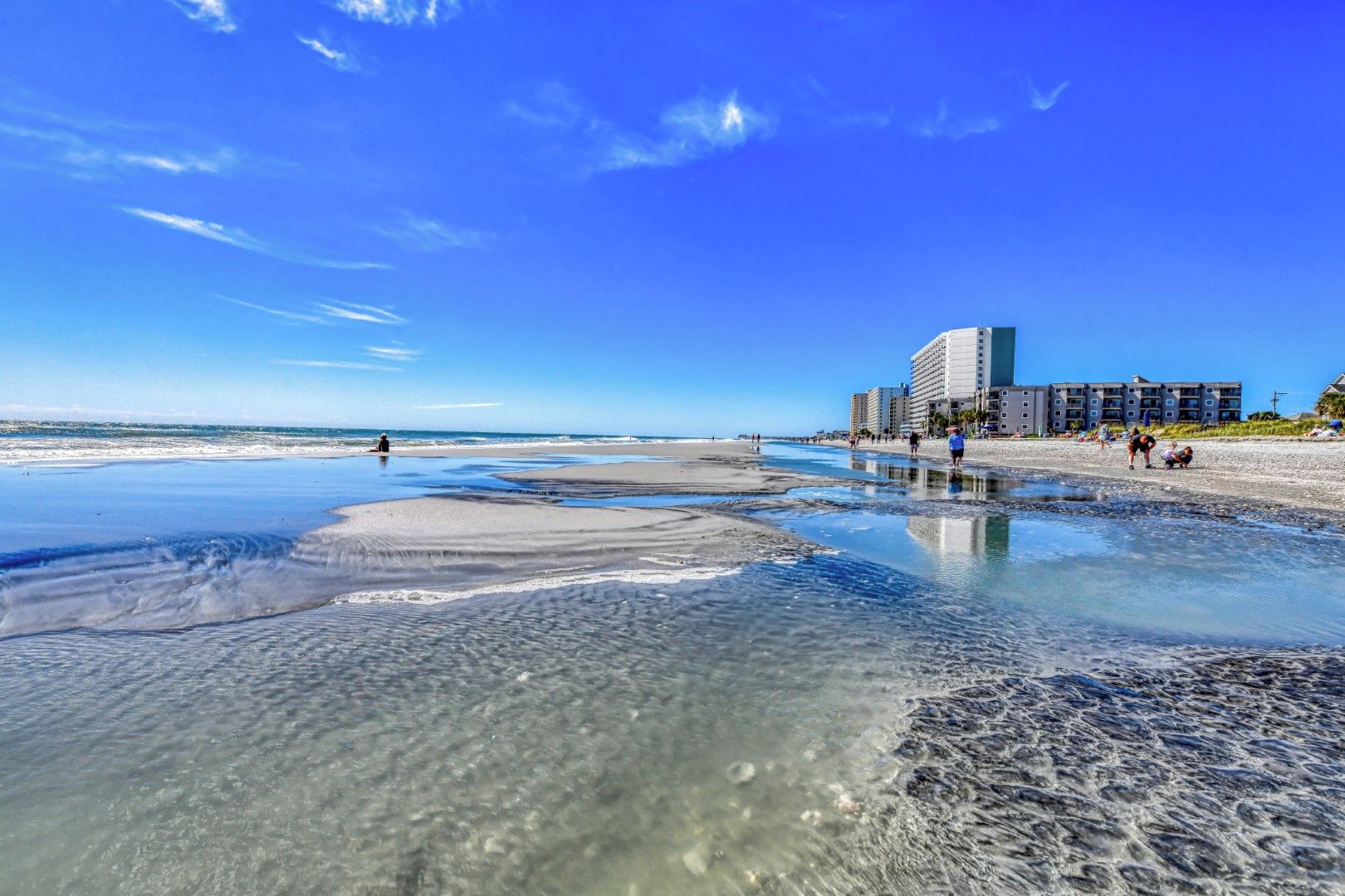A beach with clear water, a sandy shore, and a hotel in the background.