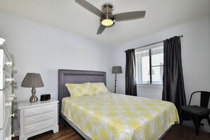 Une chambre à coucher comprenant un lit, une table de chevet, une lampe, une chaise, un ventilateur de plafond et une fenêtre avec des rideaux.