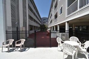 Um pátio murado com cadeiras e mesas de plástico branco, um portão vermelho e um edifício com varandas.