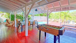 Ein überdachter Außenbereich mit einem Kicker, Holzstühlen und einem Pool im Hintergrund.