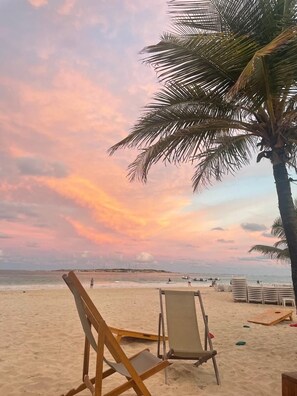A beach scene with a sunset, palm trees, and beach chairs. - Villa Soualiga orient Bay (Saint Martin)