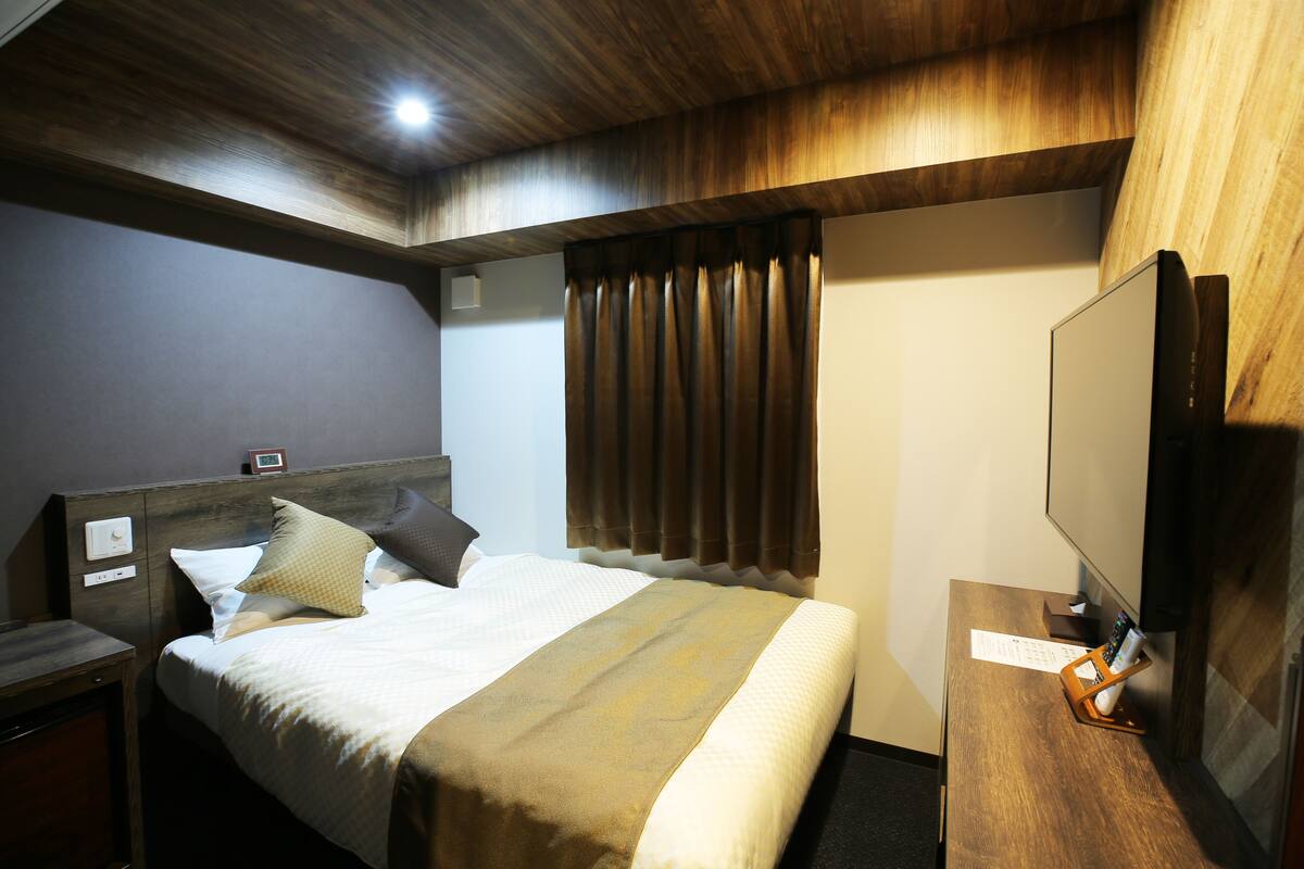 Ein Hotelzimmer mit Holzboden, einem Bett mit beiger Bettdecke und Kissen, einem Nachttisch, einem Fernseher auf einem Holzbalken und einem Fenster mit Vorhängen.