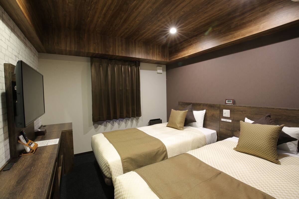 Ein Hotelzimmer mit zwei Betten, einem Flachbildfernseher, einem Holztisch und einem Fenster mit Vorhängen.