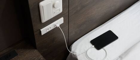 A bed with a smartphone charging on it, connected to a power outlet on the wall.