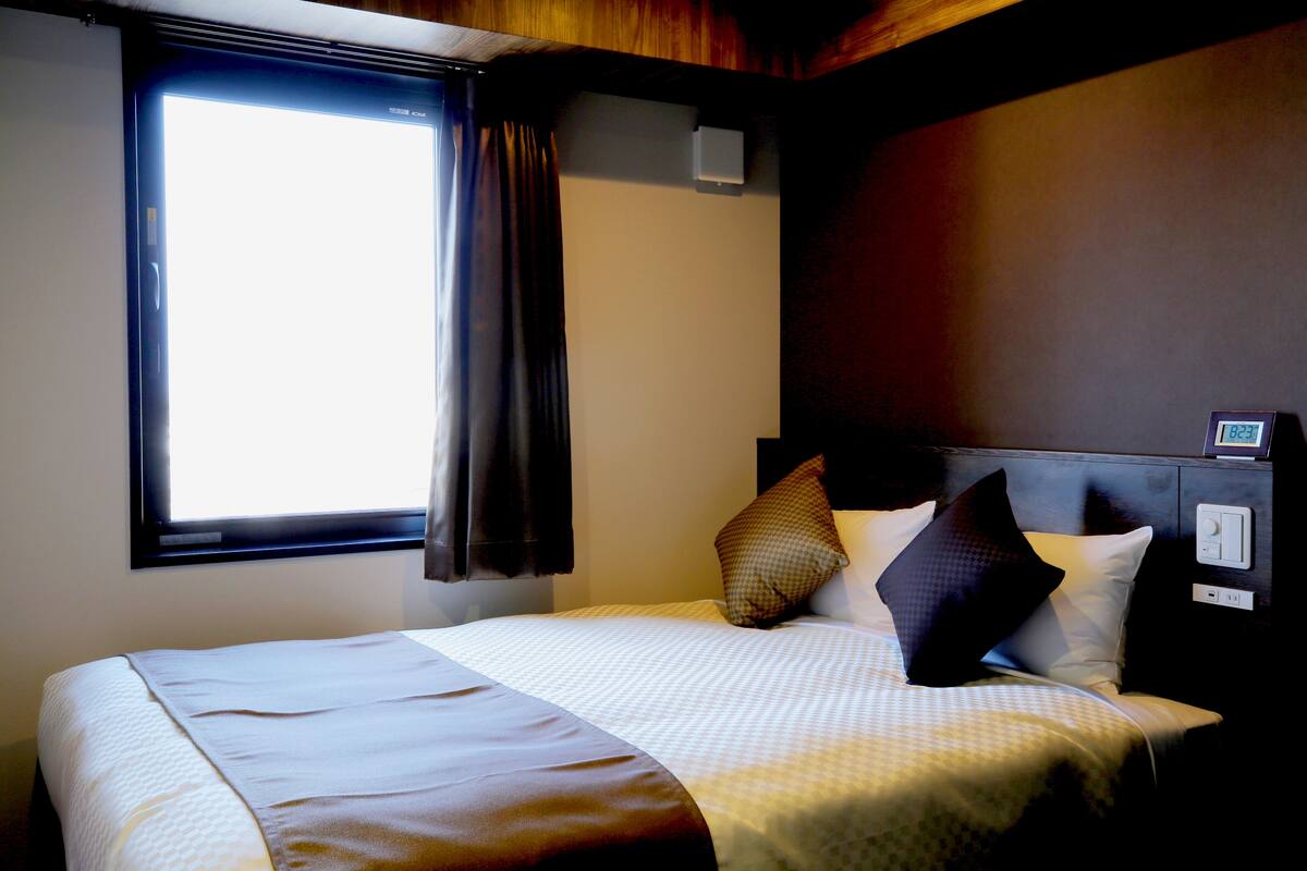 Ein Hotelzimmer mit einem großen Bett, einem Fenster mit Vorhängen, einem Nachttisch mit einer Uhr und einem an der Wand montierten Lichtschalter.