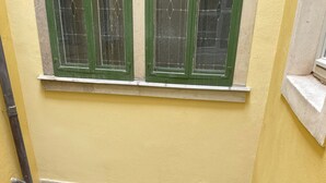 Een smalle trap die leidt naar een deur met een raam erboven, een klein raam met een houten luik, en een muur met een structuurreliëf.