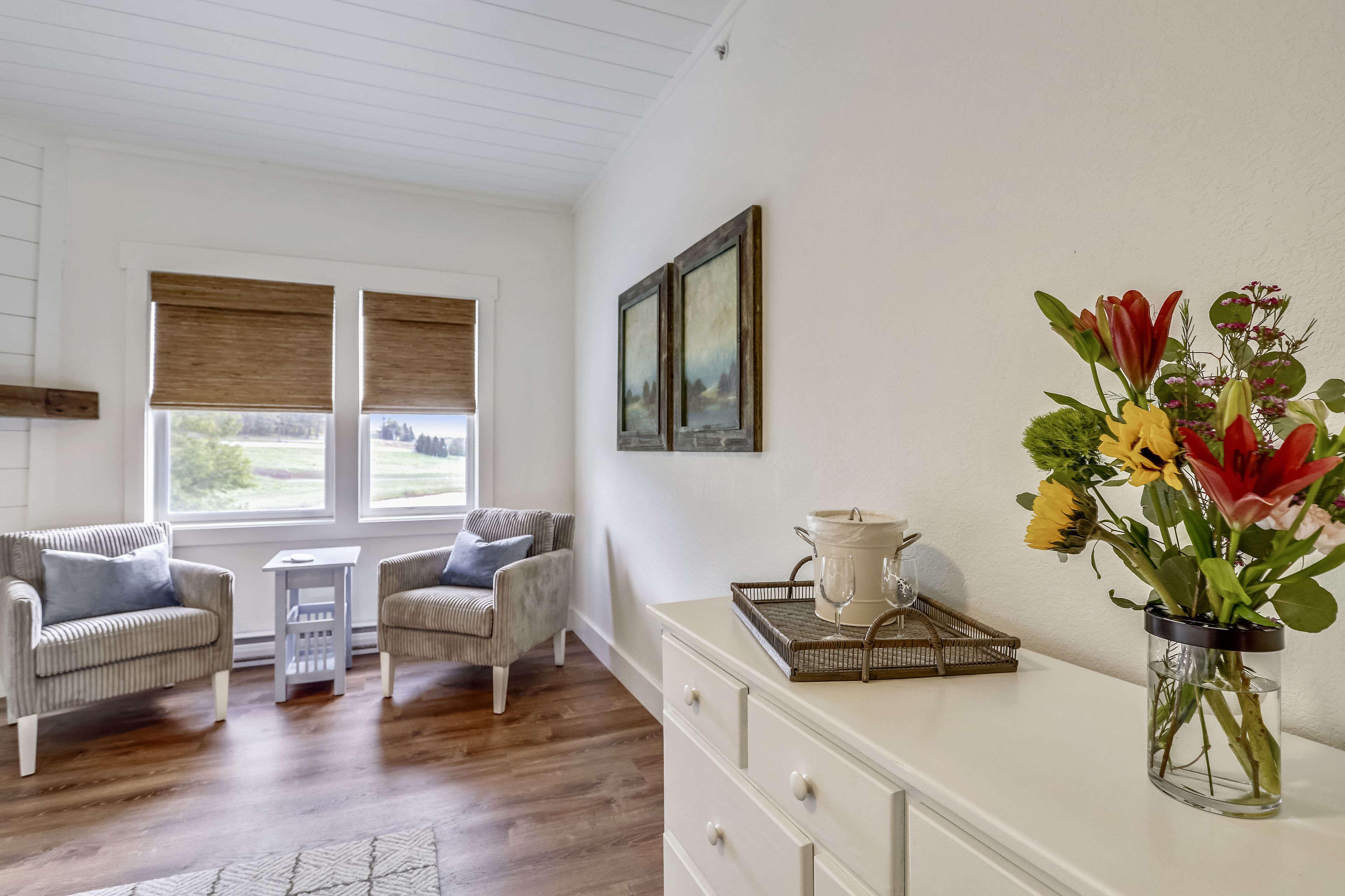 A room with two wicker chairs, a small table, a white dresser, and a vase of flowers.