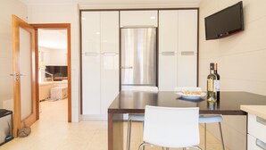 Ett modernt kök med ett kylskåp i rostfritt stål, en platt-TV som är monterad på väggen och en matplats med en vit stol och en tallrik mat.