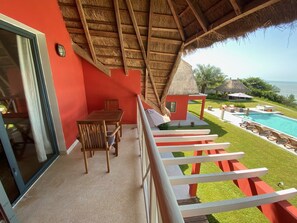 A balcony with a thatched roof, a dining area with wooden furniture, and a view of a swimming pool and ocean.