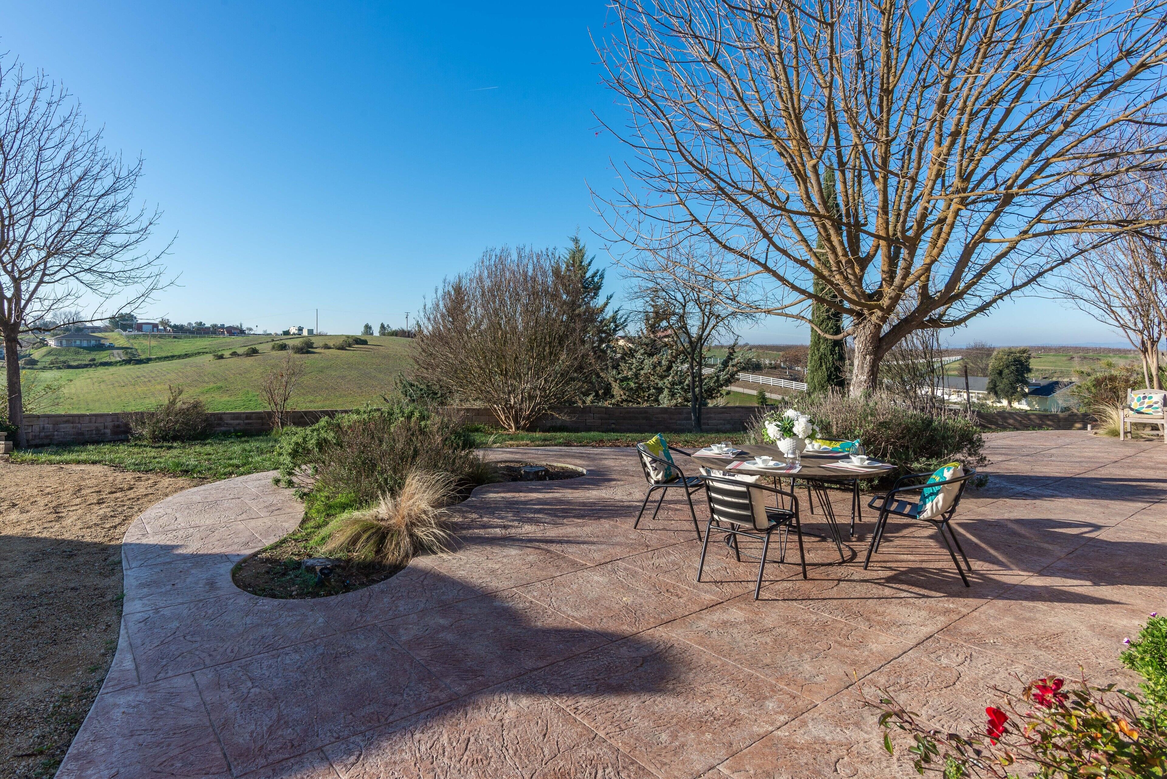 Un patio con mesa y sillas de piedra, rodeado de un jardín ajardinado y vistas al campo.