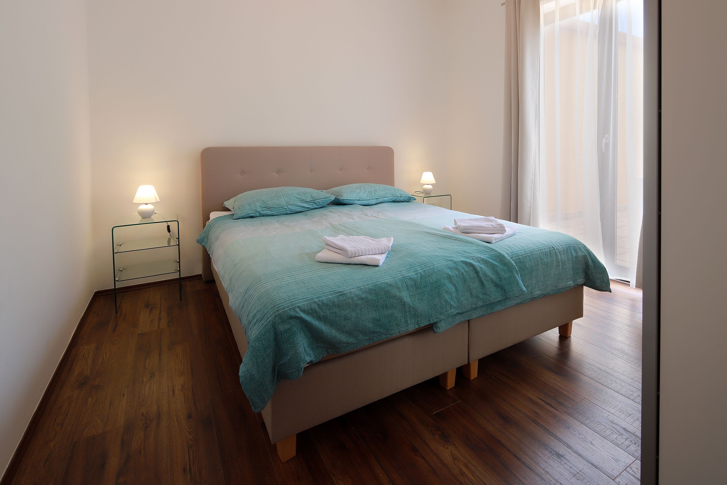Un lit bien fait, recouvert d’une courtepointe turquoise, avec deux oreillers et des serviettes pliées, le tout posé sur un plancher en bois.