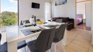 A modern dining area with a glass table, grey upholstered chairs, and a view of the outdoors. - Apartments Galboka (Mali Losinj)