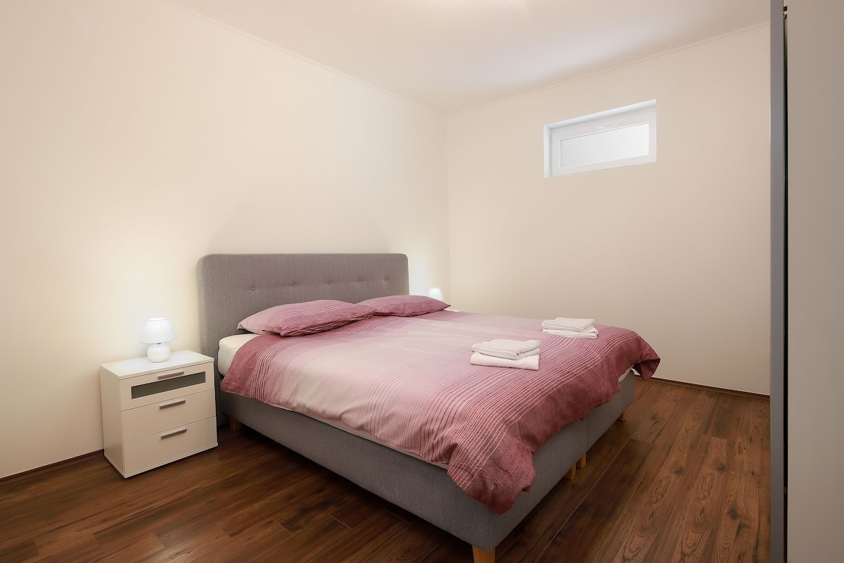 A bedroom with a bed, a nightstand, a window, and wooden flooring.