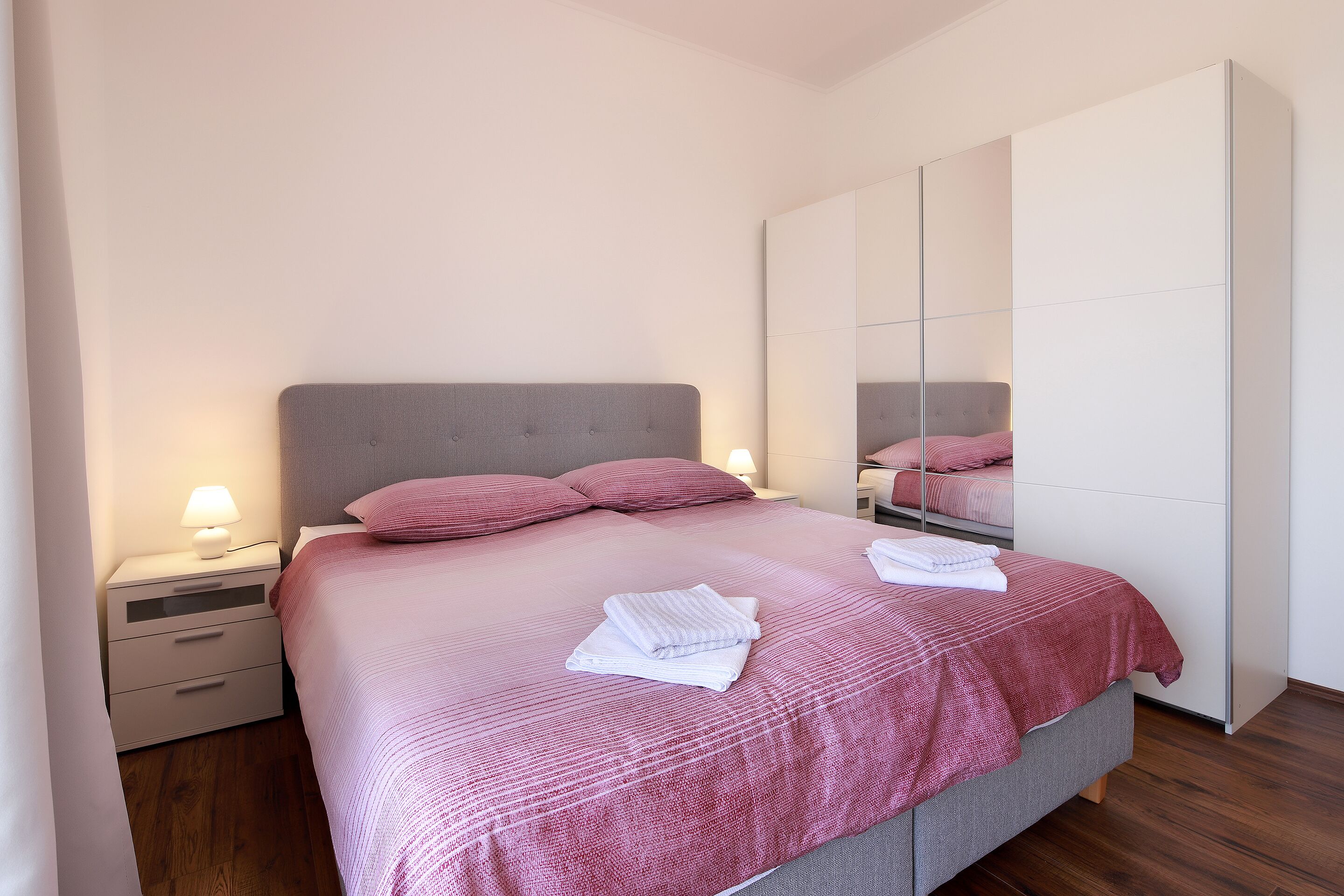 Un lit bien fait, avec une couvre-lit rose et des oreillers, une lampe de chevet et un miroir accroché au mur.
