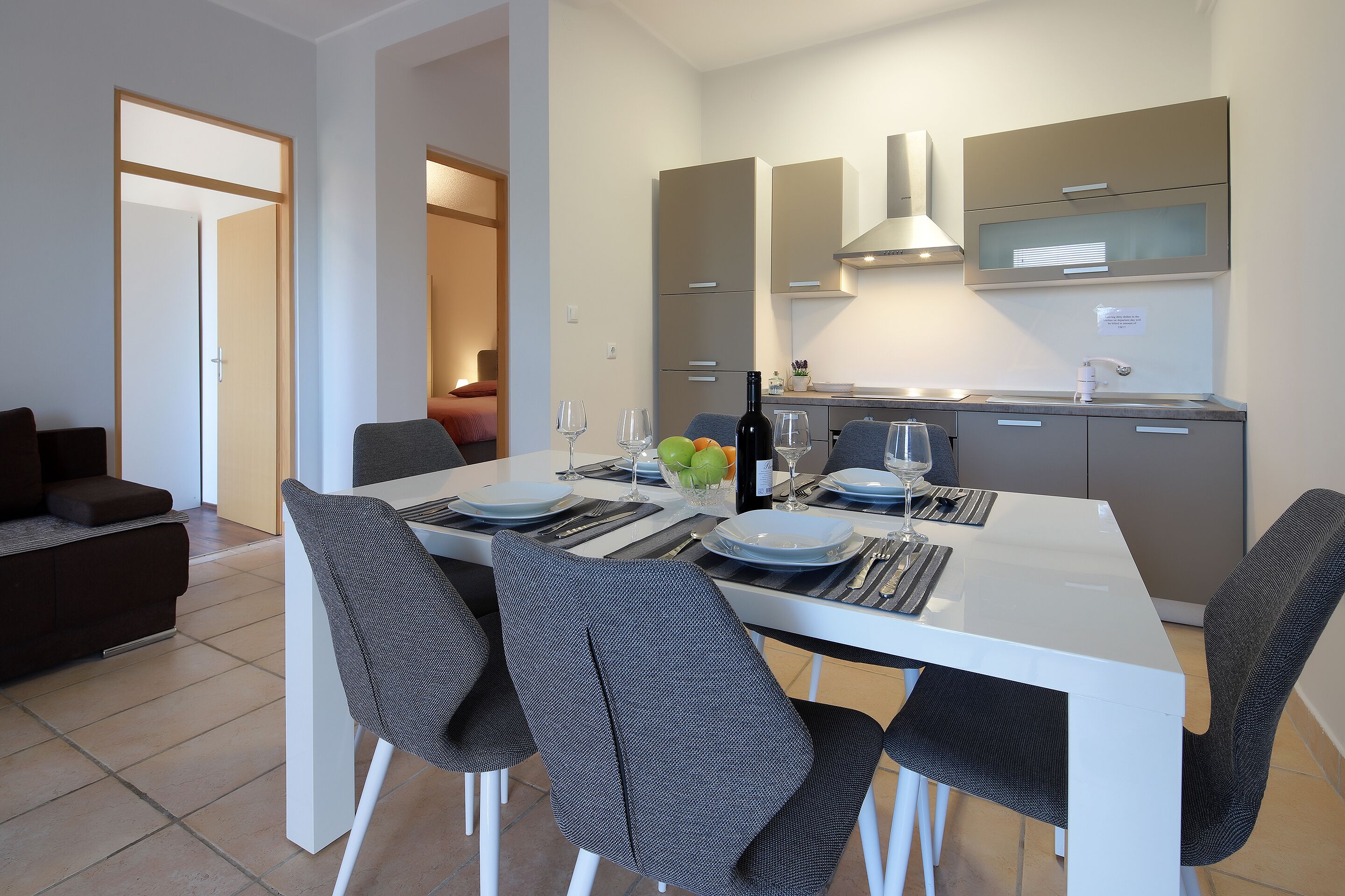 Una cocina moderna con armarios blancos, extractor de acero inoxidable y comedor con mesa blanca y sillas grises.