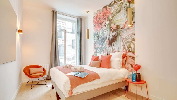 A modern bedroom with a large bed, a window with curtains, a vibrant floral wall art, and a small orange chair. - Sweett | Turenne I - Three Bedroom Apartment, Sleeps 6 (Paris)