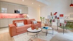 A modern living room with a pink sofa, two round coffee tables, and a kitchen area with white cabinets and a pink backsplash. - Sweett | Turenne I - Three Bedroom Apartment, Sleeps 6 (Paris)