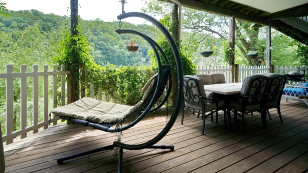Une terrasse extérieure couverte, dotée d’un fauteuil suspendu, d’une table avec des chaises et offrant une vue sur une colline boisée.
