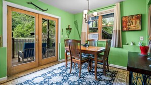 Un espace repas avec une table et des chaises en bois, un tapis à motifs, un lustre et une vue sur une terrasse et la verdure environnante.