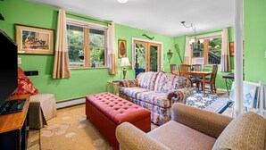 Un salon aux murs verts, avec un canapé à motifs, un pouf rouge, une table à manger entourée de chaises et une fenêtre agrémentée de rideaux.