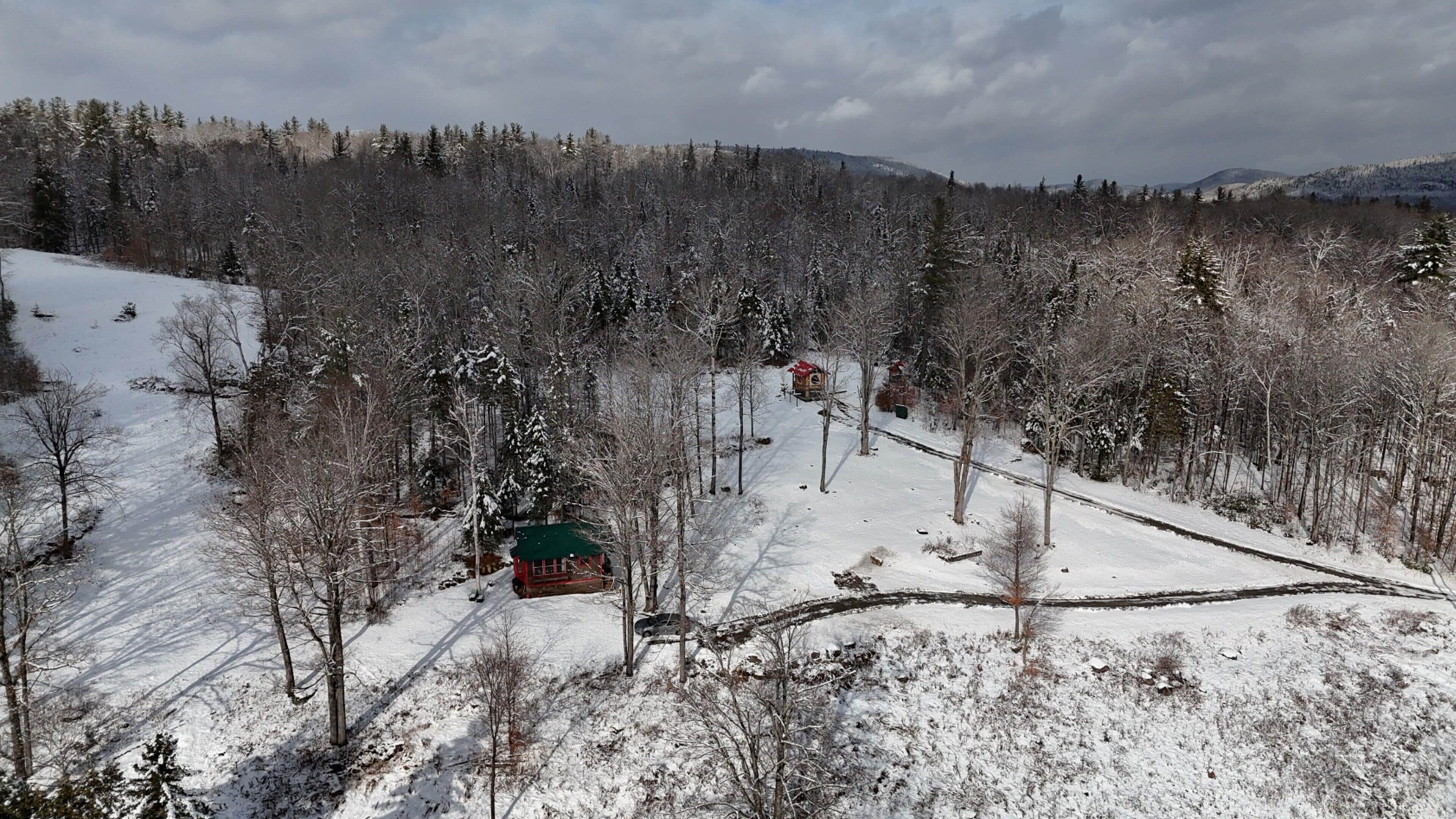 A snow-covered landscape with a cabin and a small building, surrounded by trees and a distant mountain range.