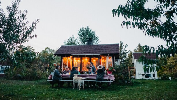 Un espace de restauration extérieur rustique, avec une table et des bancs en bois, entouré de verdure et d’un petit bâtiment doté de grandes fenêtres.