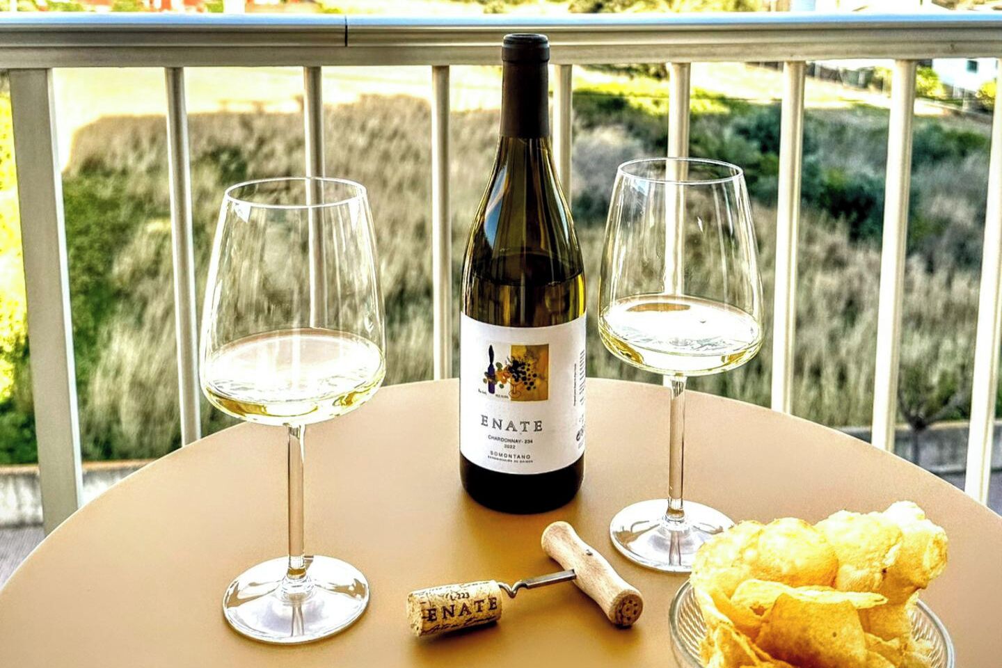 A bottle of wine, two glasses, a corkscrew, and a bowl of chips on a table with a view of greenery.