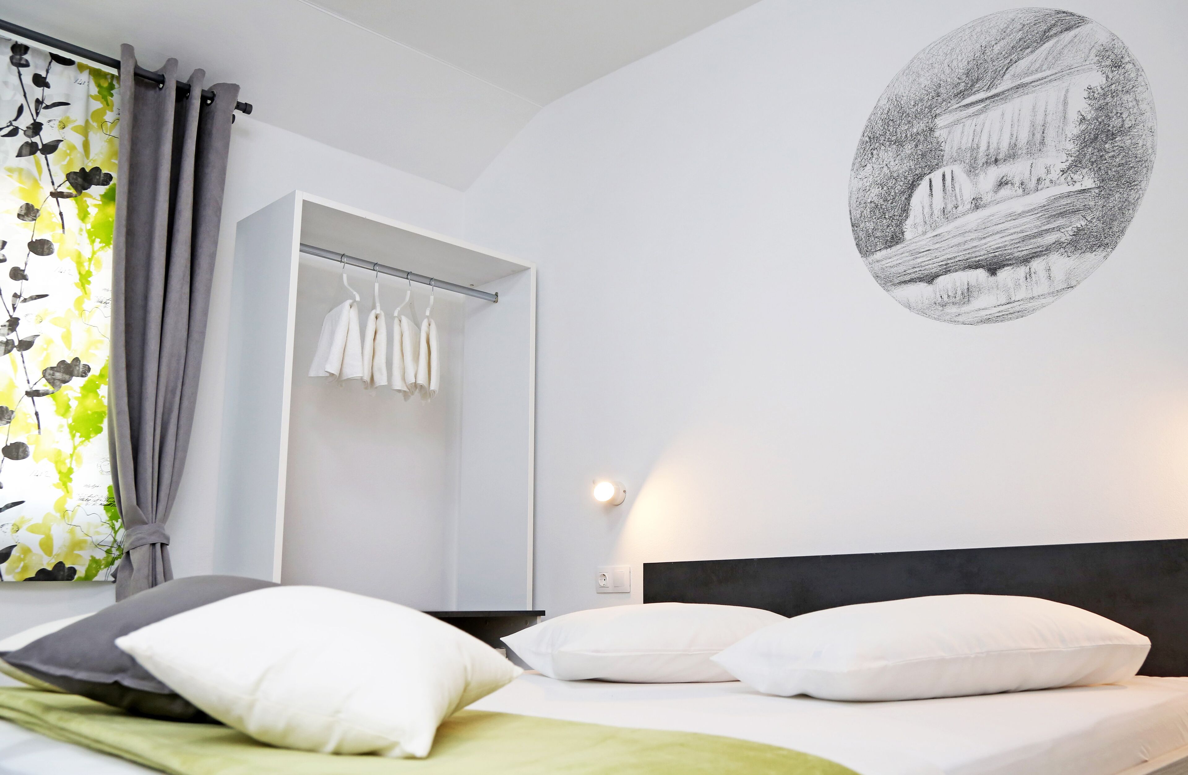 A hotel room with a bed, white pillows, a green blanket, a wardrobe, and a wall-mounted light.