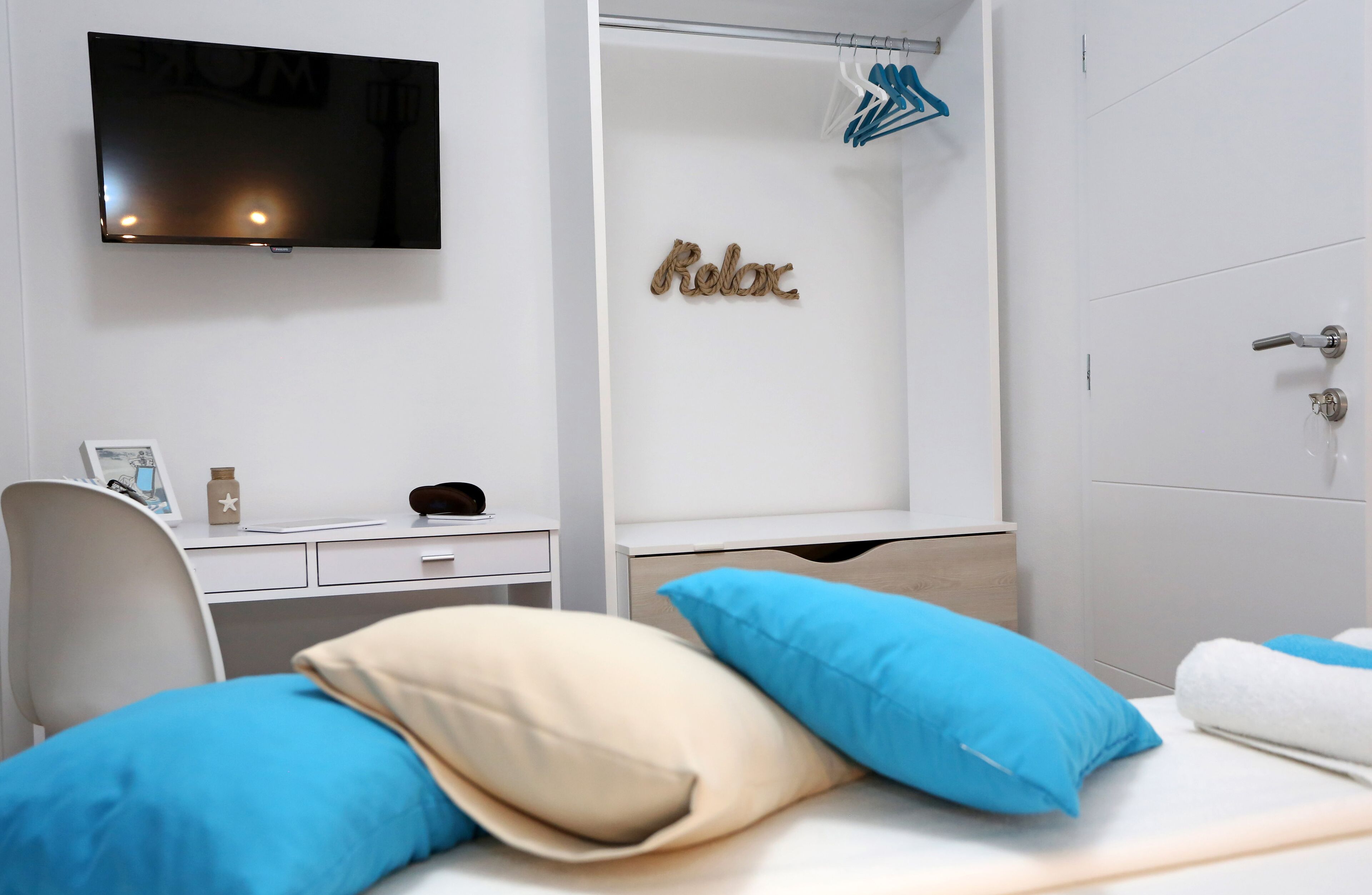 Una habitación de hotel moderna con una televisión de pantalla plana, un escritorio con ordenador, una cama con almohadas azules y beige, y una zona de ducha con perchero.
