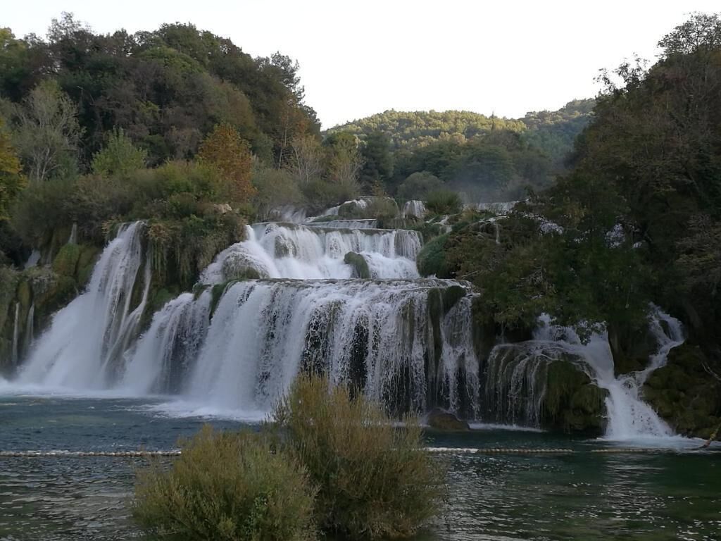 A waterfall cascading over multiple tiers into a calm river surrounded by dense forest.