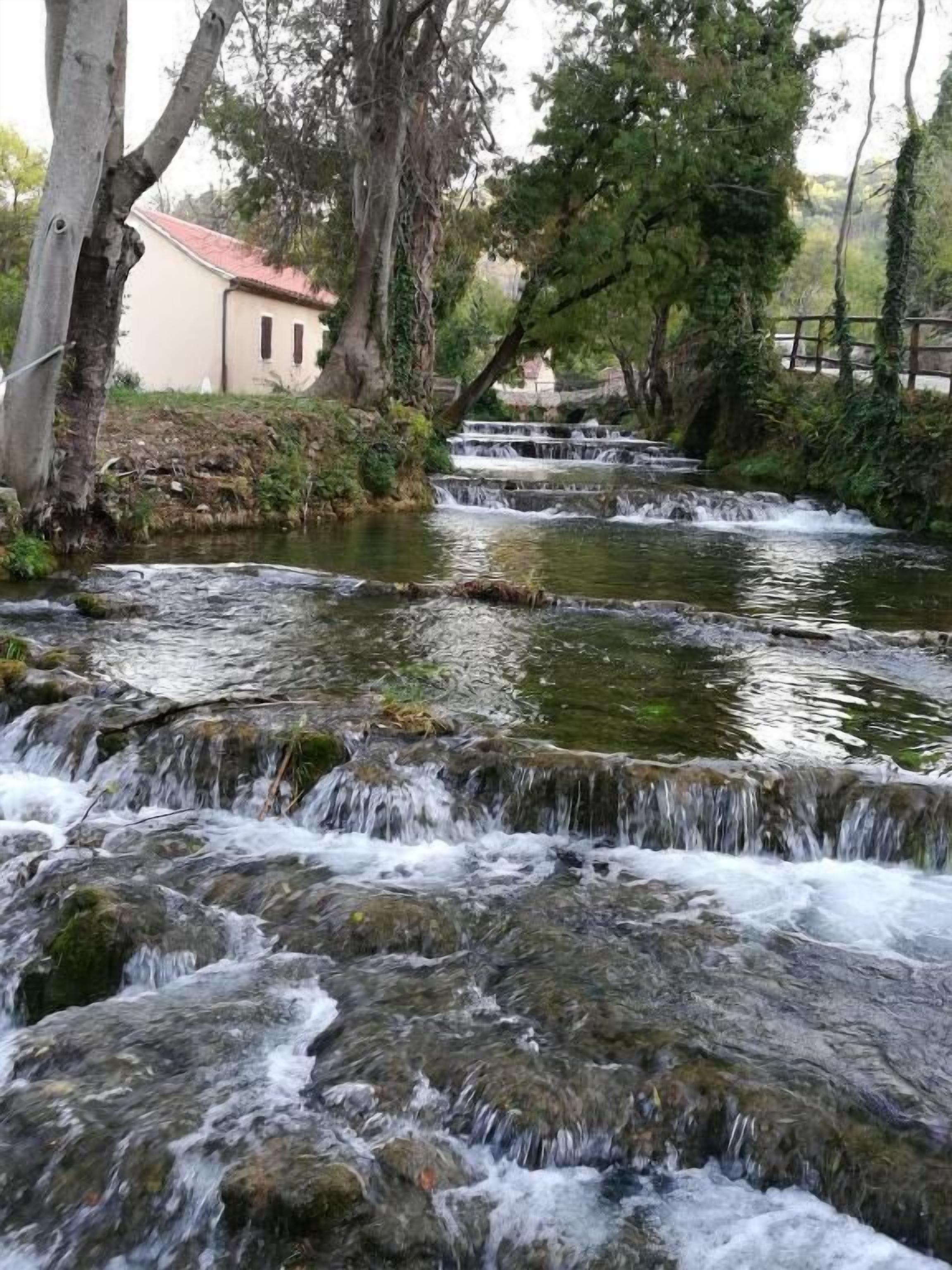 A natural water feature with cascading water over rocks, surrounded by trees and a building in the background.