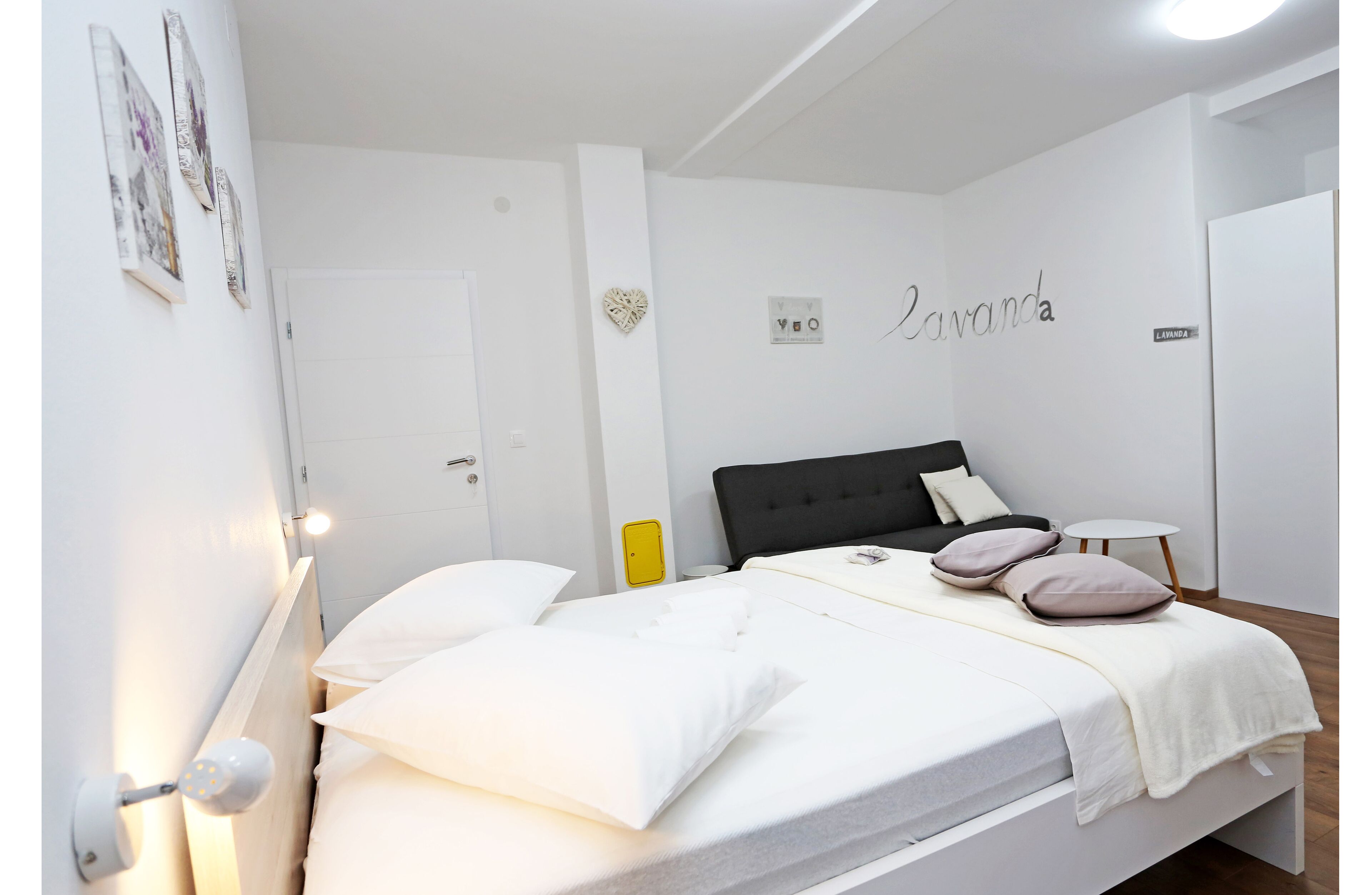 Una cama bien hecha con ropa blanca y un cabecero negro, una mesita de noche con una lámpara, y una pared decorada con un adorno en forma de corazón y la palabra 'Lavanda'.