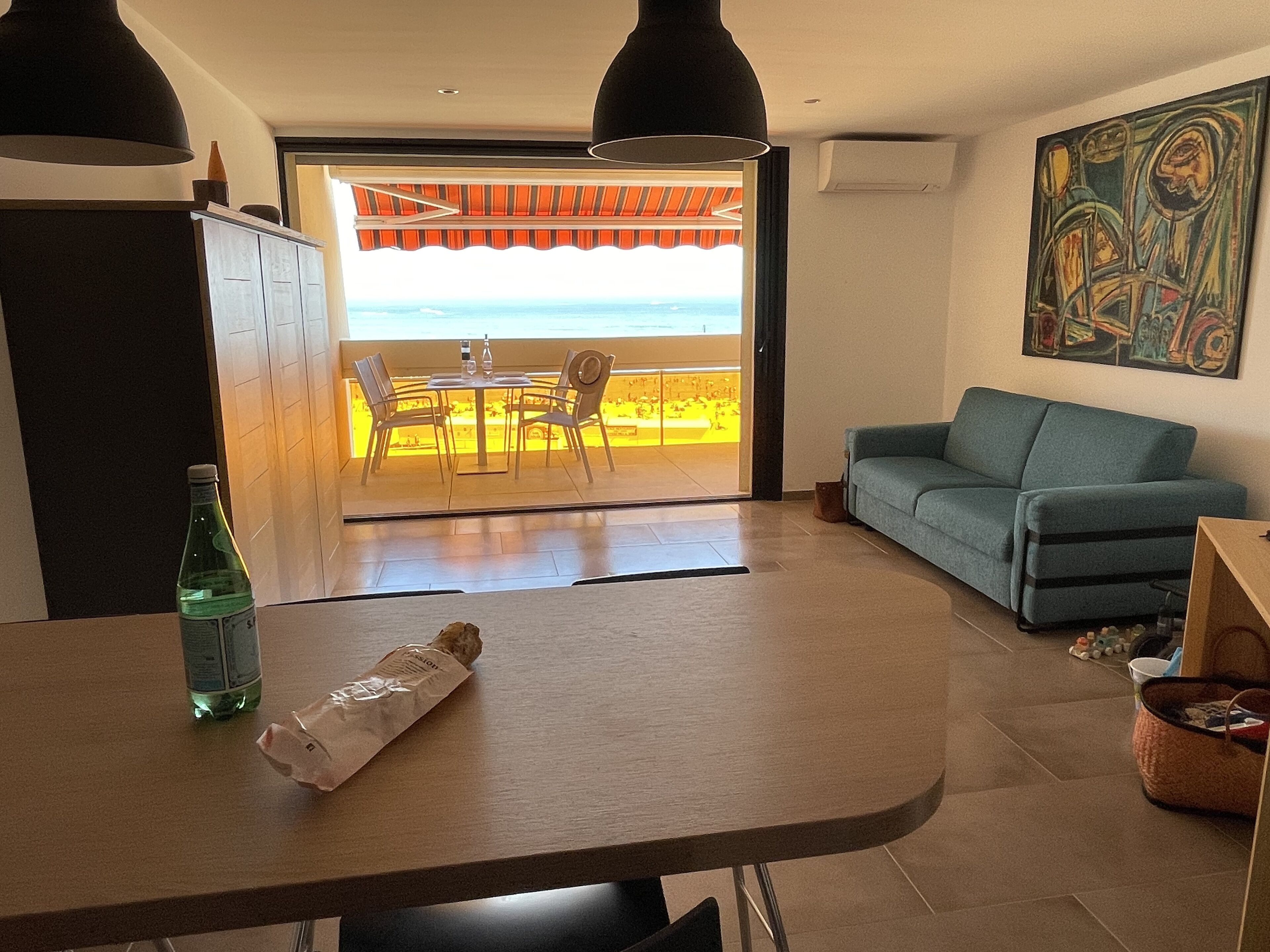 A room with a view of the ocean, a dining table with chairs, a sofa, and a painting on the wall.