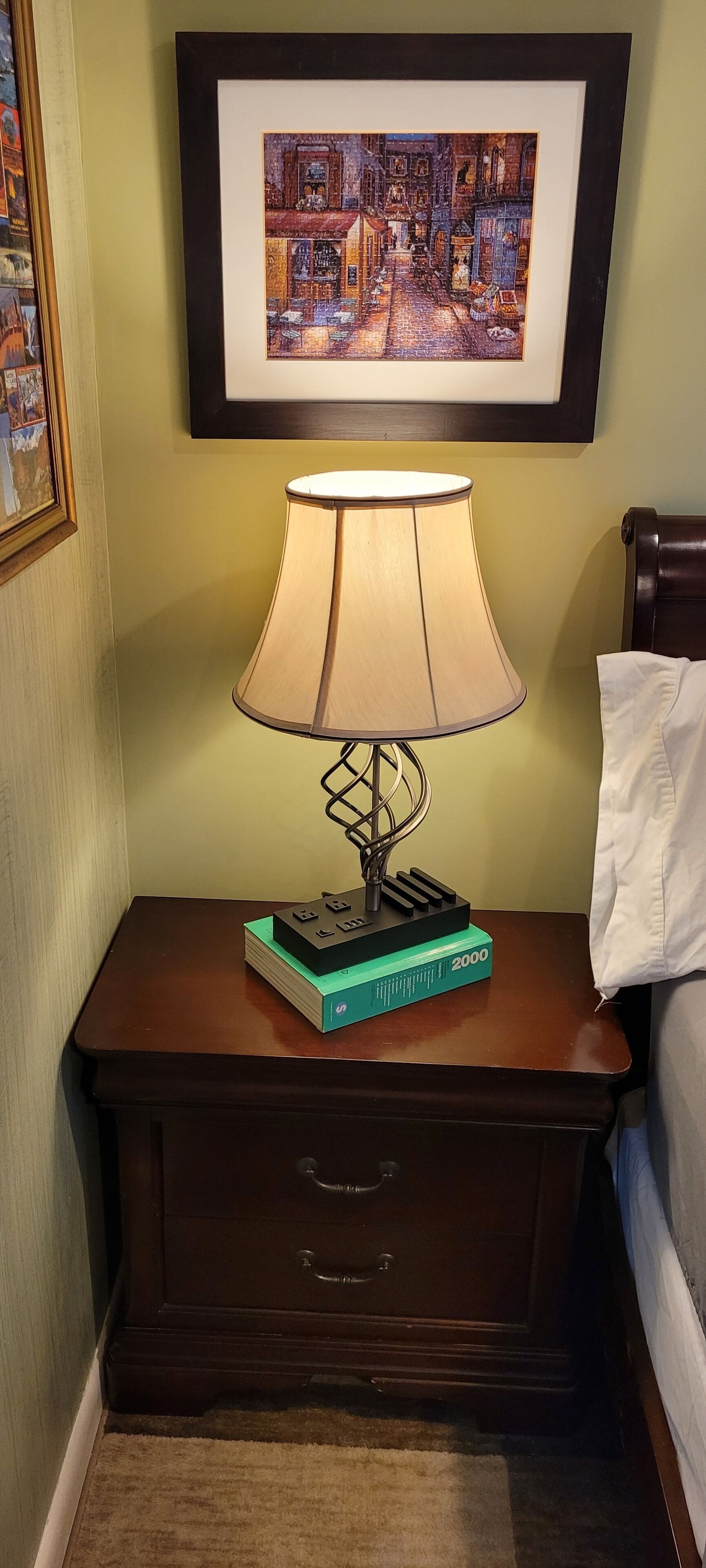 A bedside table with a lamp, a framed picture above it, and a book on the table.