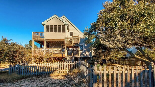 A two-story house with a balcony, surrounded by a wooden fence and trees. - June 27-July 4th is OPEN. CHECK THE DISCOUNTED RATES. (Corolla)