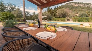 An outdoor dining area with a wooden table set for four, surrounded by wicker chairs, with a pool and mountains in the background. - Finca Gloria for 4 persons with pool (Pollensa)