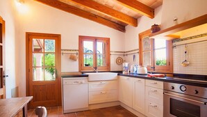 A kitchen with wooden beams, white cabinets, a dishwasher, a stove, and a window with a view of greenery. - Finca Gloria for 4 persons with pool (Pollensa)